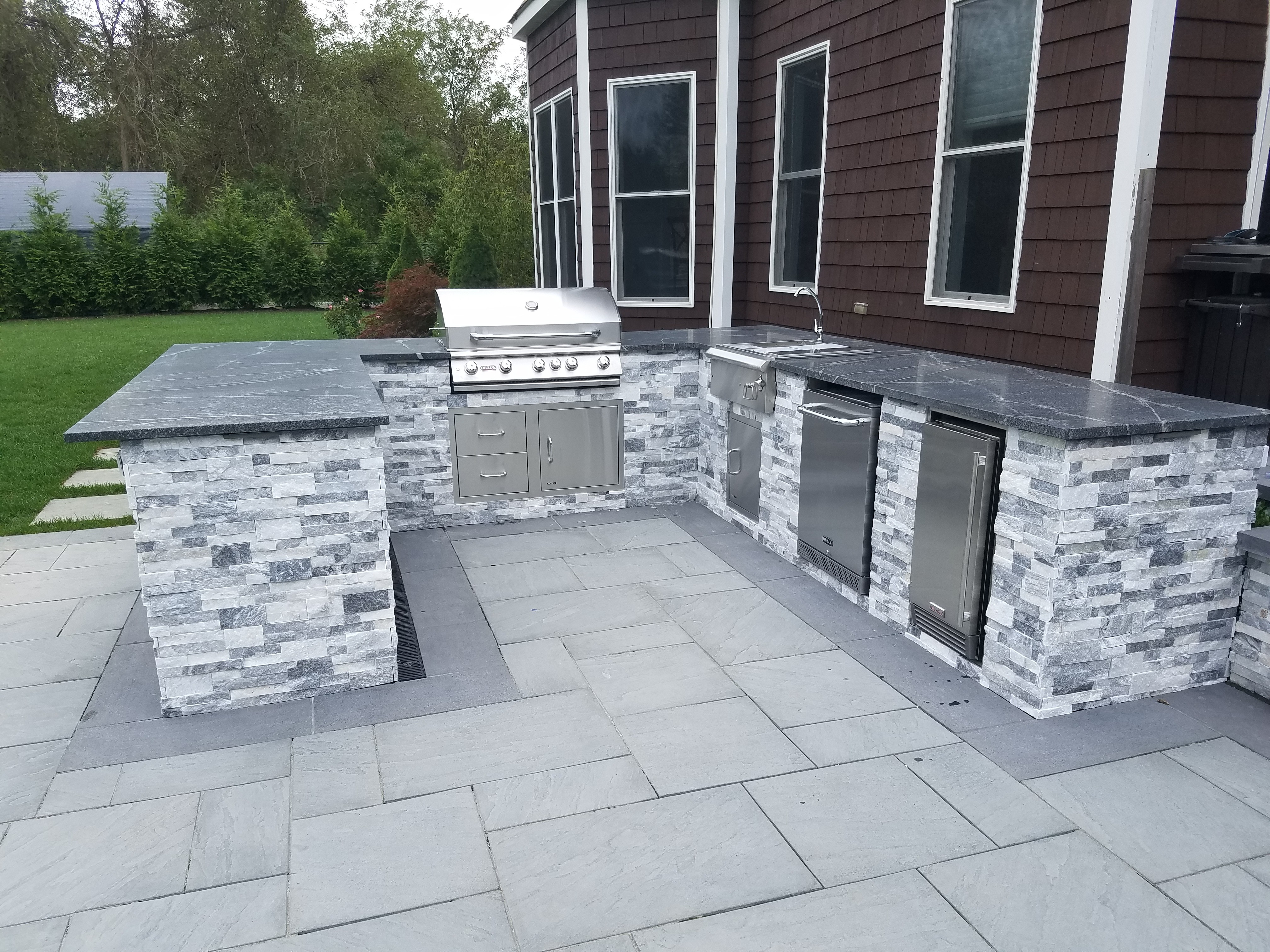 Modern gray-stone outdoor kitchen with built-in grill, sink, and fridge