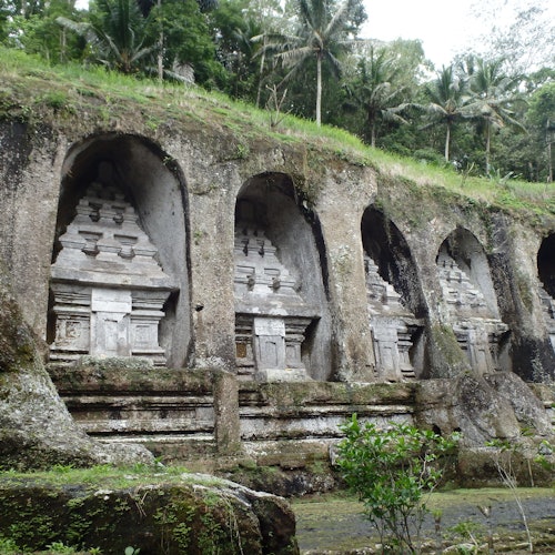 石刻的寺廟神龕鑲嵌在岩石坡地中，四周環繞著綠色植物和棕櫚樹。