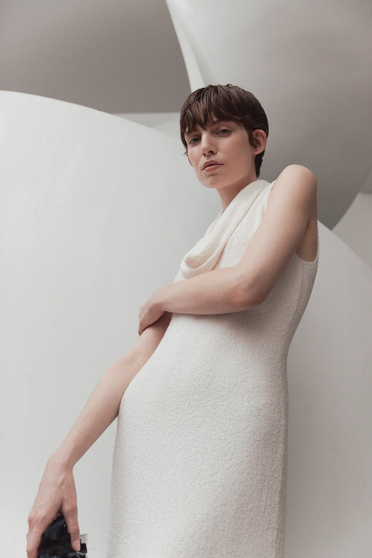 Full-length view of the Aida Barni SS26 collection white textured knit maxi dress with a draped neckline worn by a model on a white staircase.