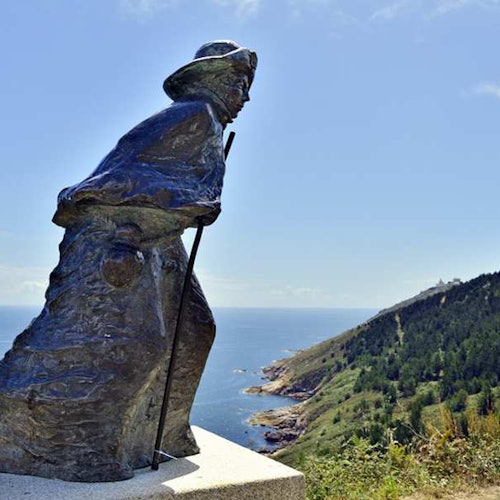 Statua in bronzo di un escursionista con un bastone da passeggio, che domina un paesaggio costiero con alberi e scogliere sotto un cielo azzurro e limpido.