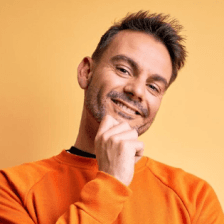 A person with a light beard, wearing an orange sweater, is smiling with a thoughtful gesture against a warm yellow background.
