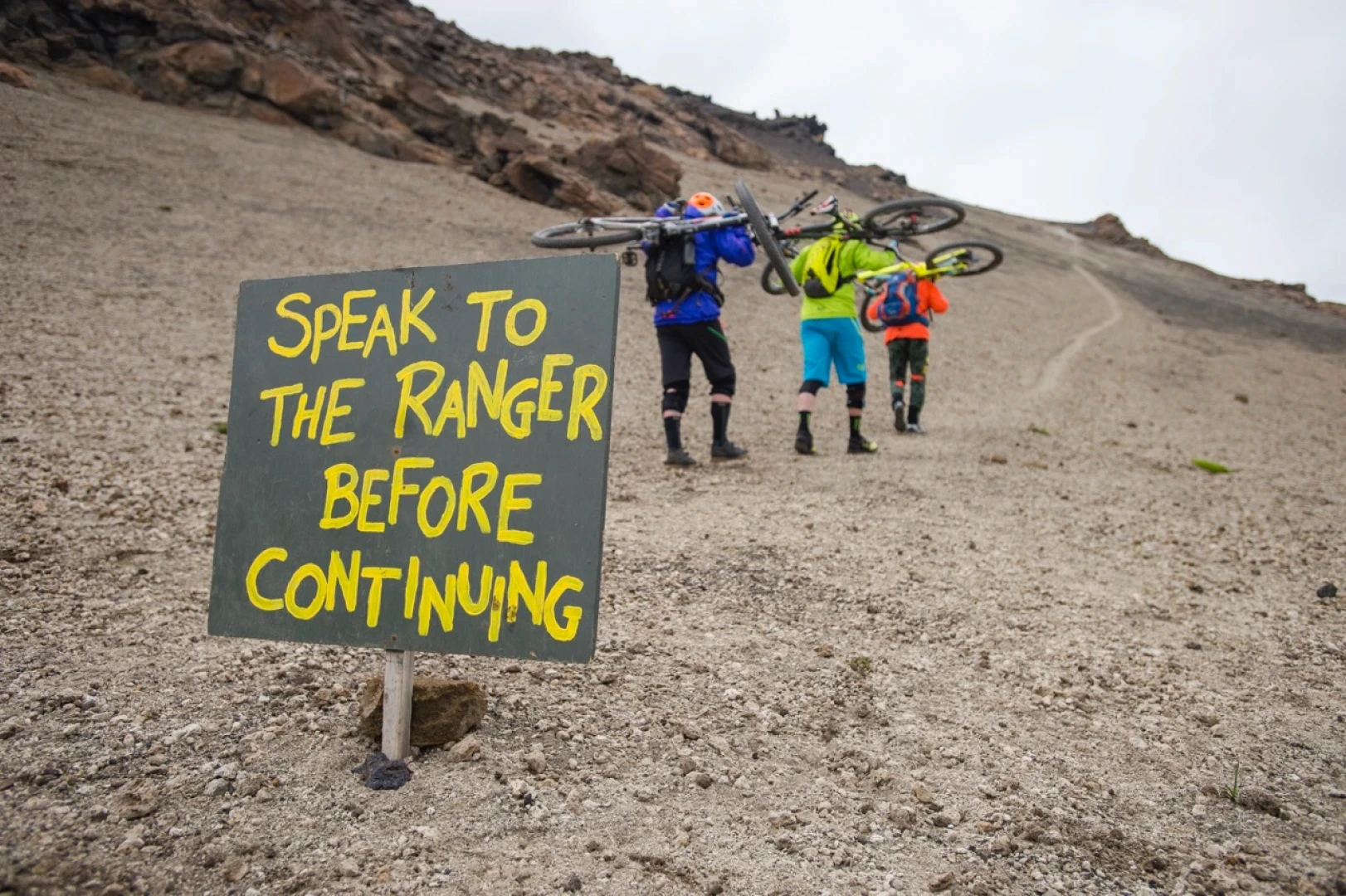 A sign in foreground and riders hike their bikes away from camera in Landmannalaugar