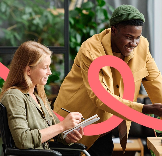  A woman in a wheelchair writing in a notebook while a man in a yellow jacket leans over to collaborate with her
