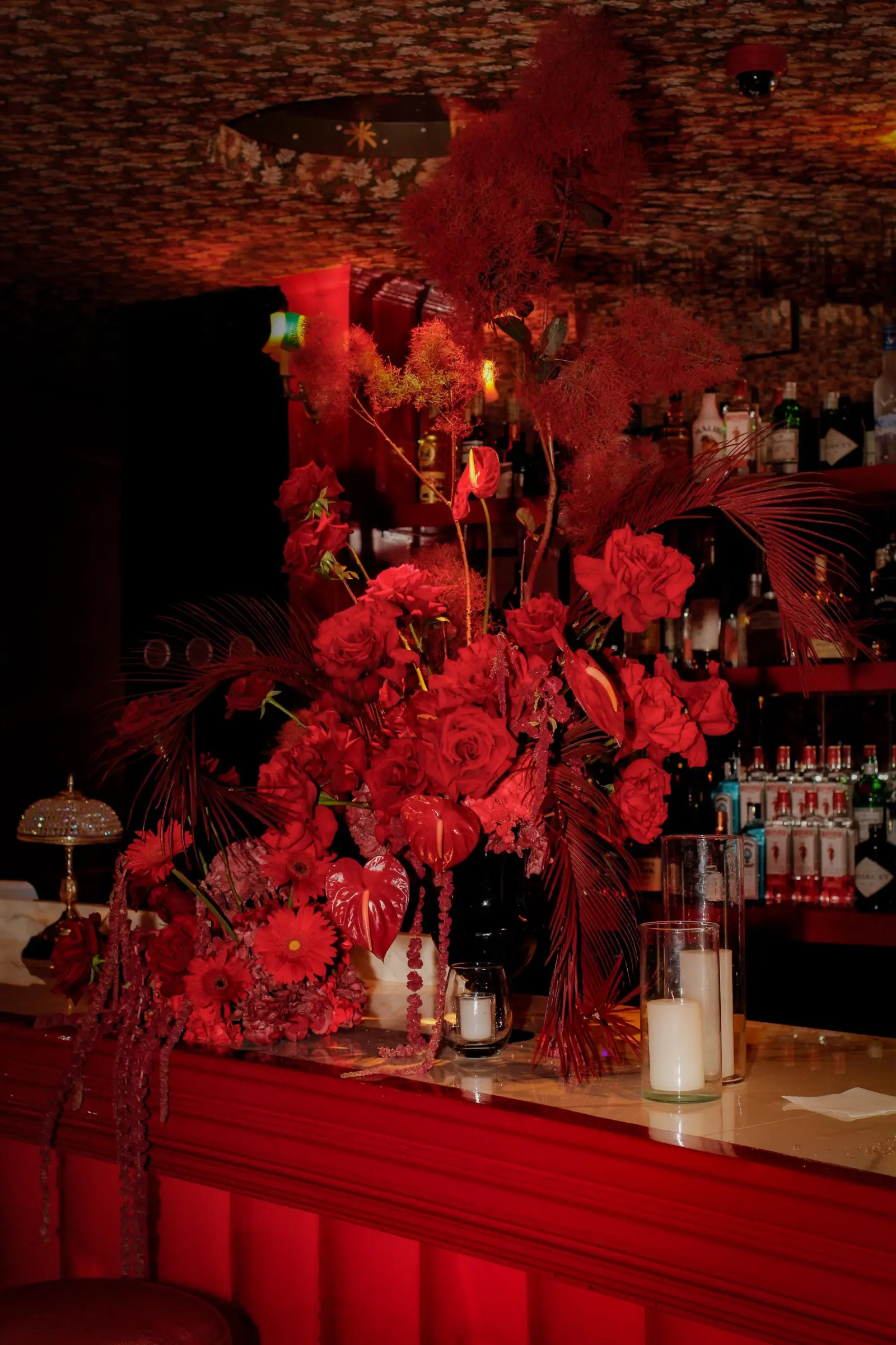 Barra de bar decorada con gran arreglo floral rojo, velas blancas y botellas de licor al fondo.
