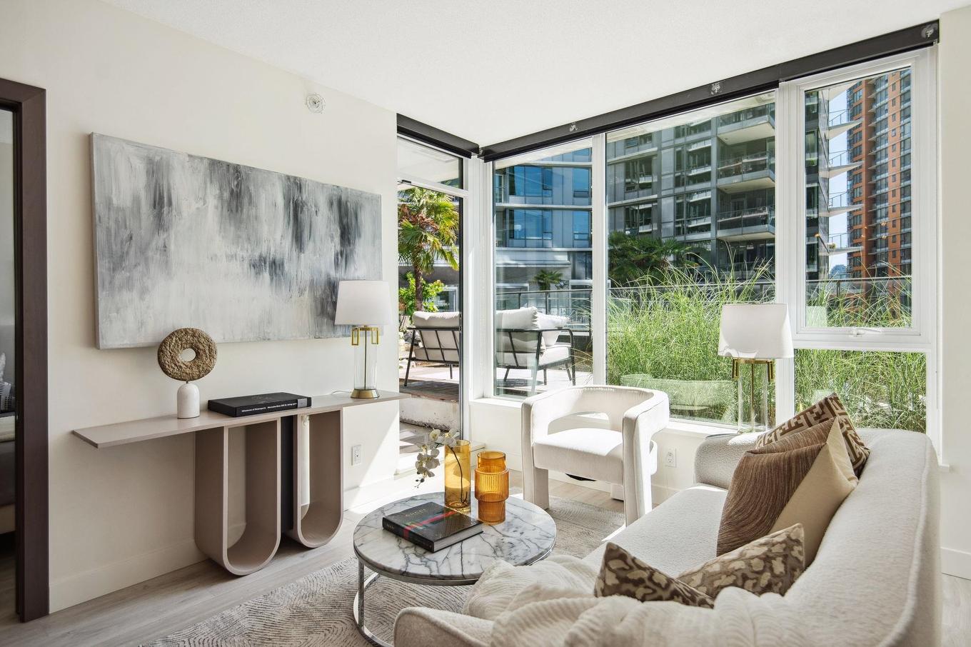 Bright and modern living room with large windows, comfortable seating, and abstract wall art. Lush greenery outside.