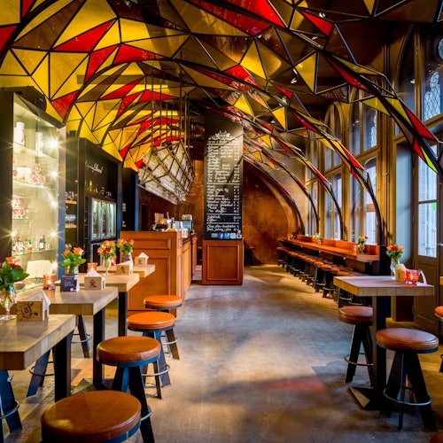 Egy stílusos kávézó színes geometrikus mennyezettel, fa asztalokkal és székekkel, egy bárpulttal, menütáblával, és virágokkal az asztalokon.