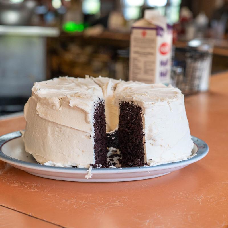 A whole homemade chocolate layer cake under thick swirled vanilla frosting, one wedge cut away to show the dark crumb inside