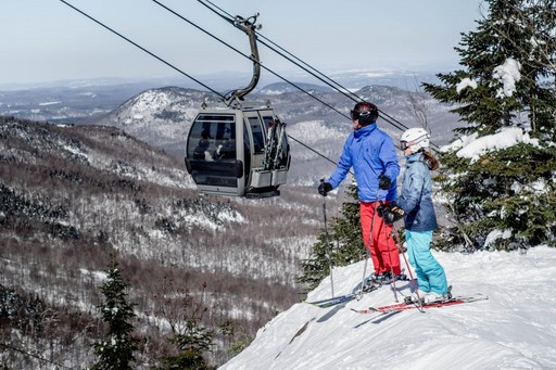 Skieurs descendant une piste de ski du Mont-Orford avec télécabine et montagnes enneigées à l’arrière-plan.