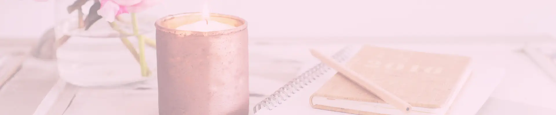 A lit candle, notebook, pencil, and vase of flowers on a rustic white desk, adding warmth to the workspace to boost focus..