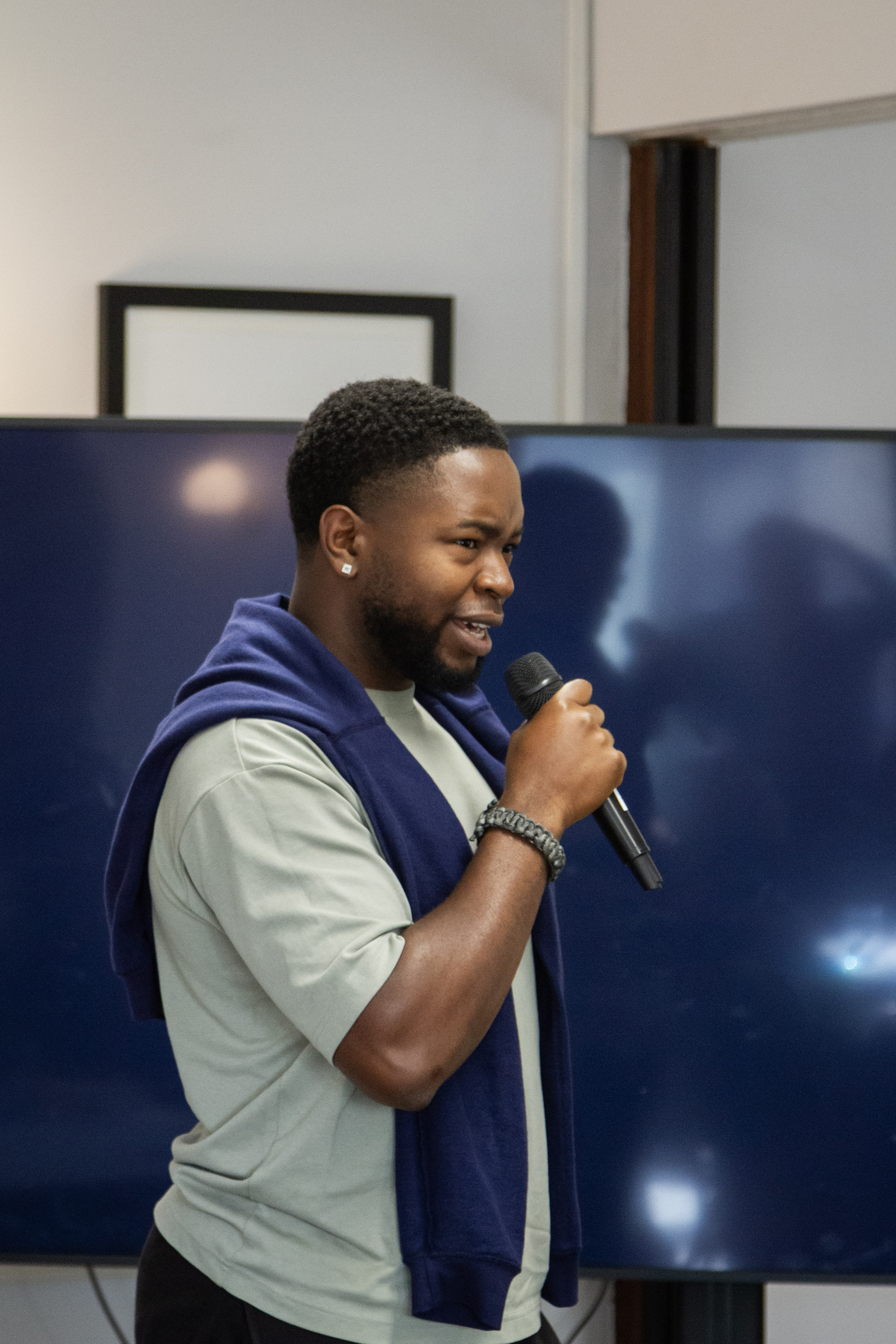Man holding a microphone and speaking, wearing a blue valuable sweater over a light shirt.