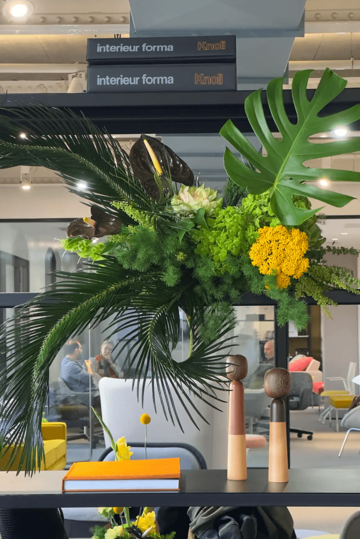 Instalación floral asimétrica con follaje verde vibrante y flores amarillas integrada en estantería de oficina.