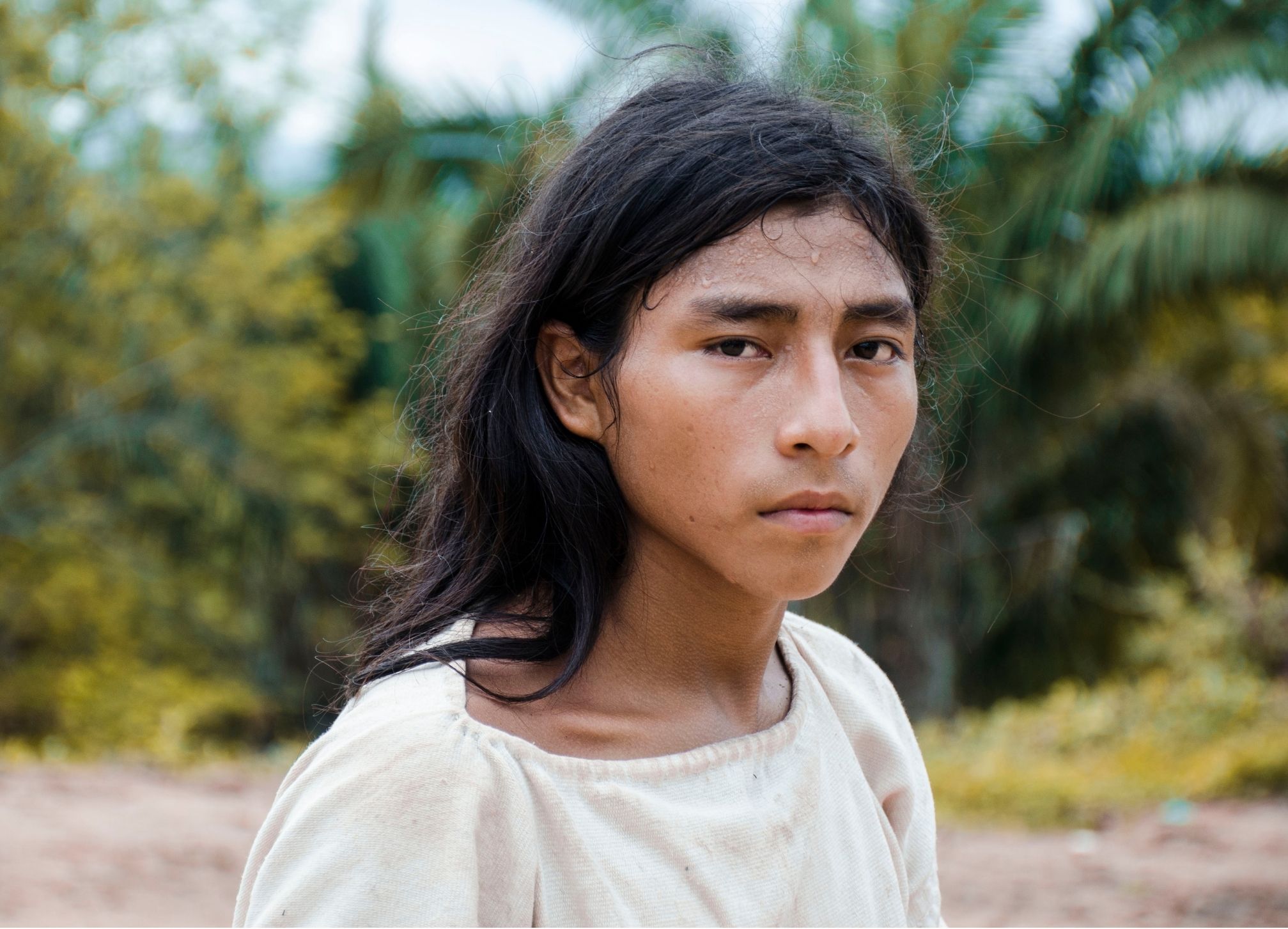 A young person with long black hair and a serious expression stands outdoors. They wear a simple, light-colored shirt and are surrounded by lush greenery. The background features trees and foliage, and the individual appears to be sweating slightly, suggesting a warm environment. The focus is on their face, which conveys a sense of intensity.