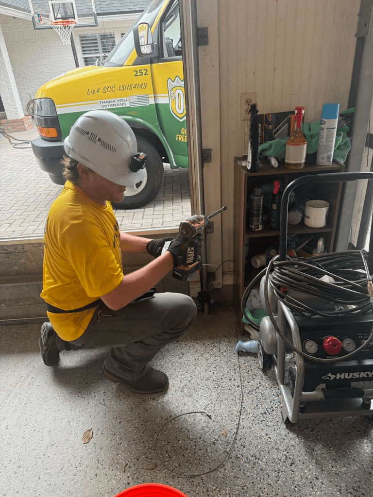 A technician in a yellow shirt and helmet uses a power drill in a garage. Nearby, a service truck is parked outside. The scene is busy and focused.