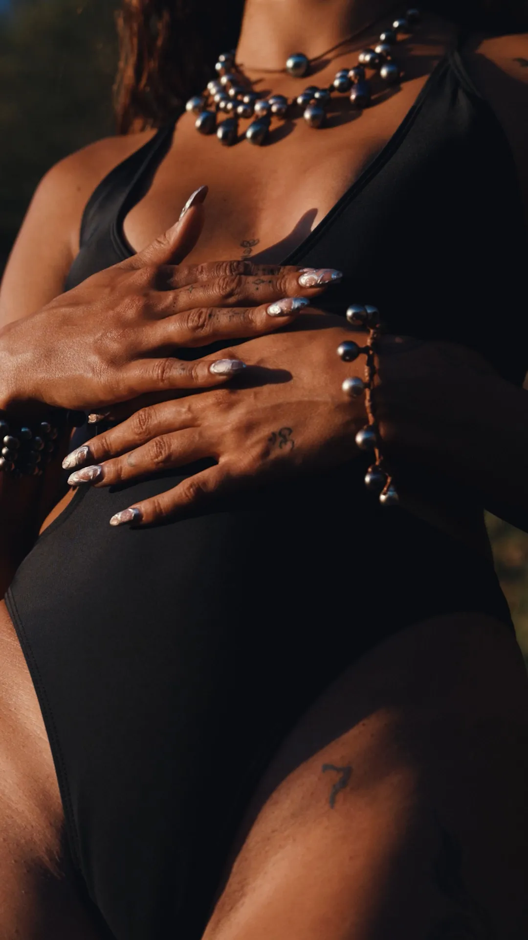 Close-up of tattooed hands resting on sun-warmed skin, adorned with a bold multi-strand Tahitian pearl necklace and a matching pearl bracelet — sensual, raw, and deeply crafted. Jewelry with a soul, crafted by hand — Manos de Oro Saint Barth.