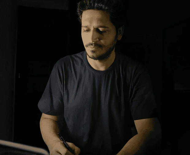 Man sitting in a dimly lit room, wearing a beige shirt and looking contemplative.