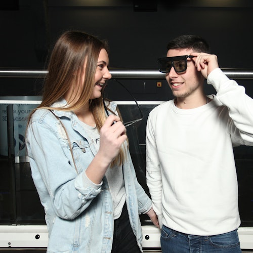 A woman and a man smiling at each other, each holding up a pair of 3D glasses. They are standing in front of a railing in a dark room.