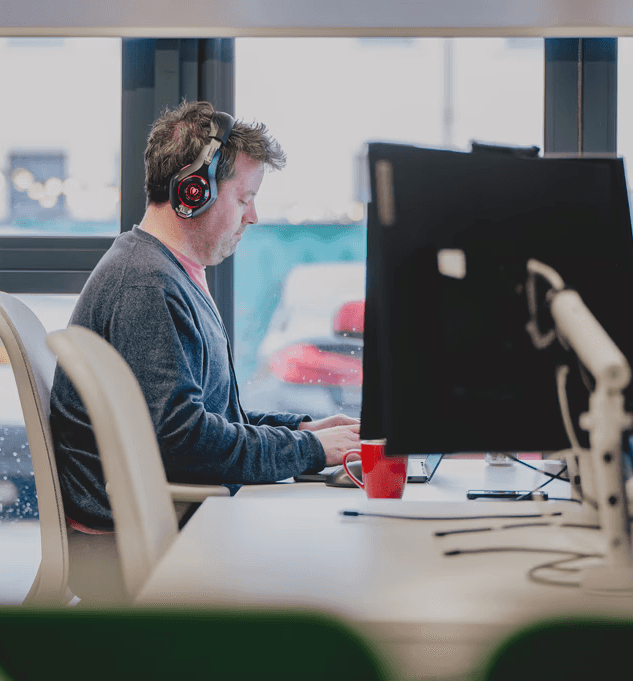 **Alt text:** Person wearing over-ear headphones works at a computer desk in an office, typing on a keyboard between large monitors, with a red mug on the desk and a window with blurred city lights in the background.