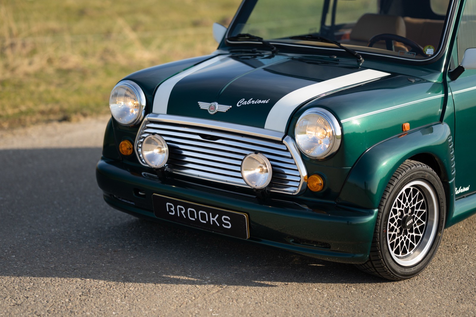 Close-up of a classic green car with white accents and headlights, parked on a road with grassy surroundings.