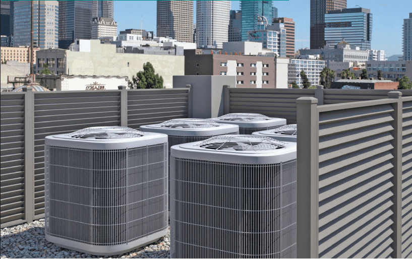 A tall, grey louvered aluminum privacy screen enclosing multiple rooftop commercial air conditioning units against a city skyline.