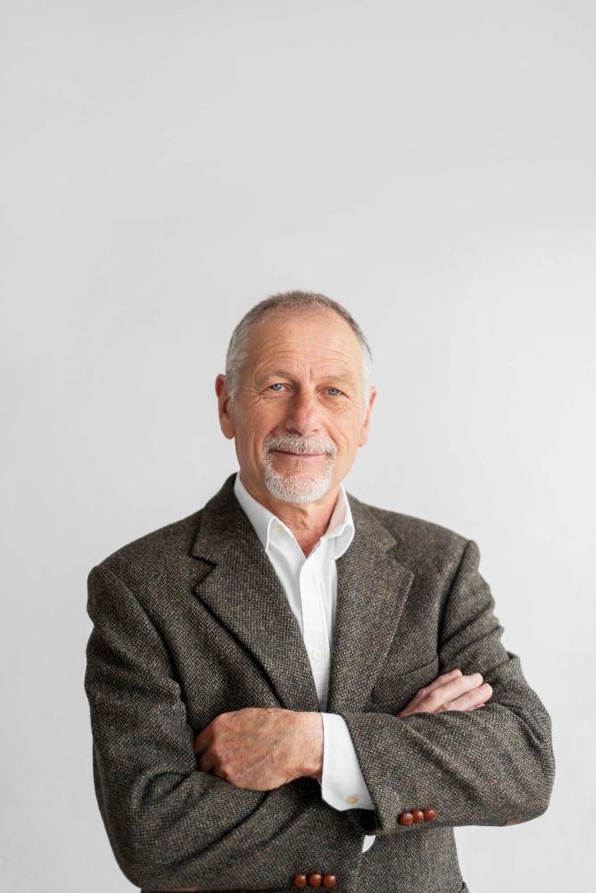 Confident senior man in a tweed blazer smiling with arms crossed against a neutral background