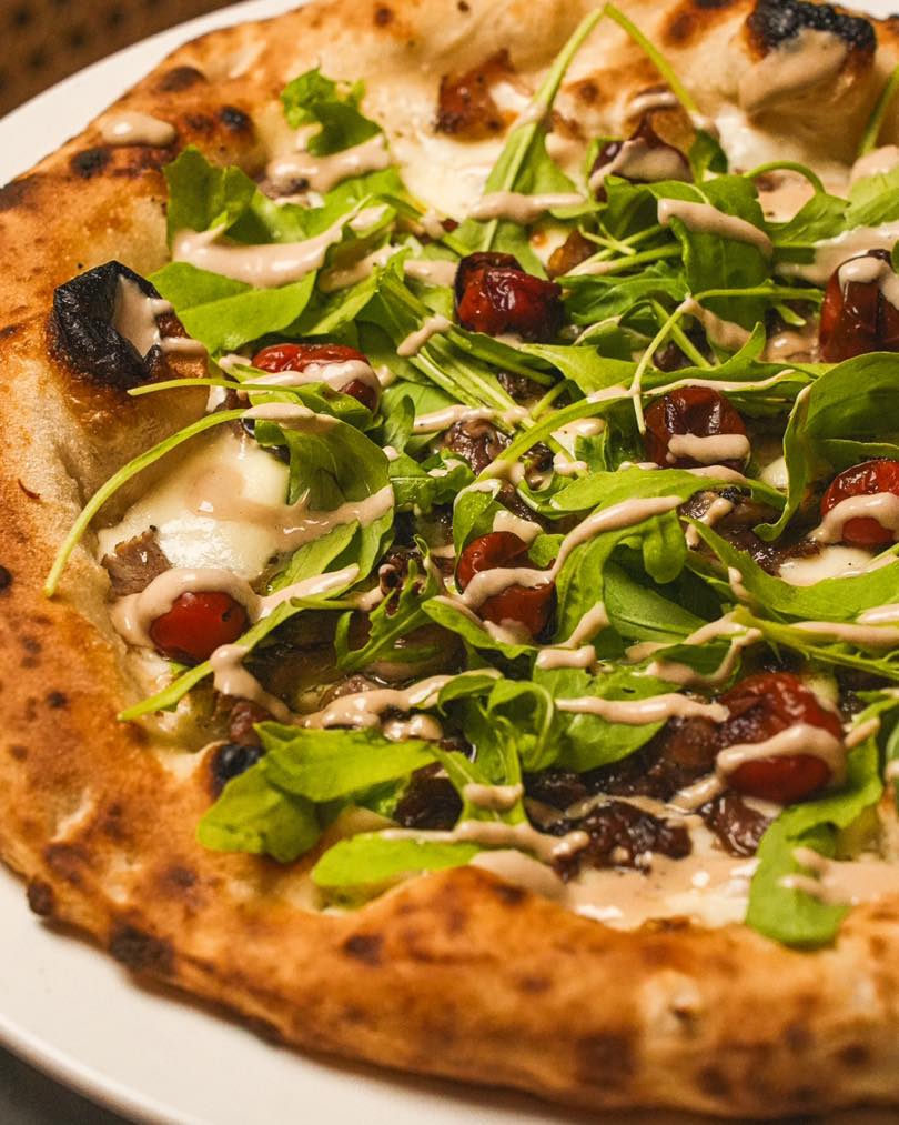 A close-up of a pizza topped with fresh arugula, roasted cherry tomatoes, and a drizzle of sauce, showcasing the crispy crust and flavorful ingredients.
