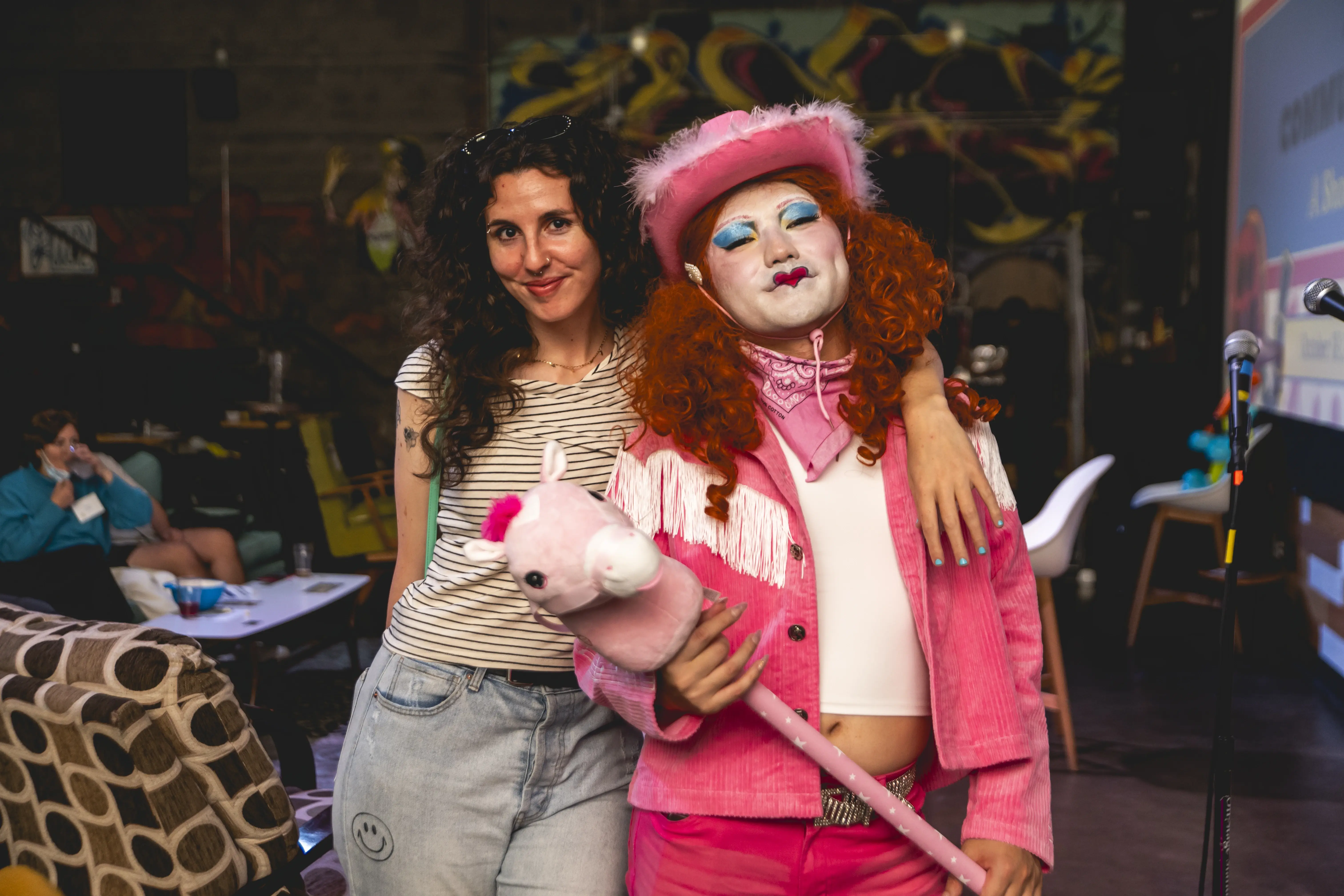 Uma pessoa com cabelo comprido e cacheado, vestindo uma camisa listrada, está de braços dados com outra pessoa vestida com um vibrante traje de cowboy rosa, completo com chapéu, pintura facial branca e segurando um brinquedo de unicórnio rosa, em um cenário interno animado.