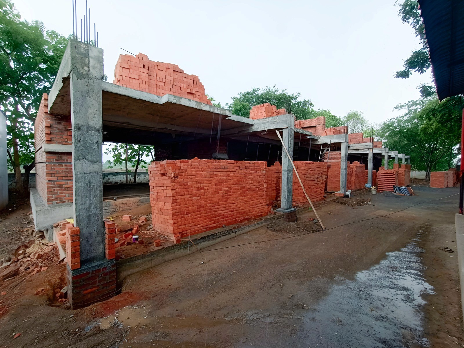 The House of Hope school under construction: concrete frames and new brick walls among trees on a wet dirt site.