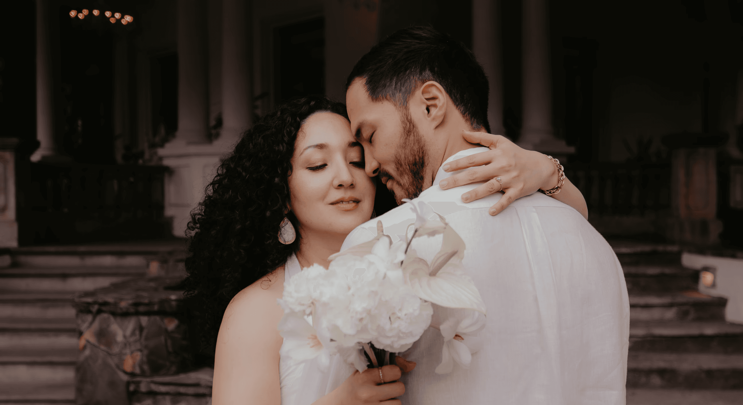 A bride and groom hold hands tenderly, the bride's bouquet of dried flowers hanging low. They stand in a softly lit, elegant interior, conveying romance.