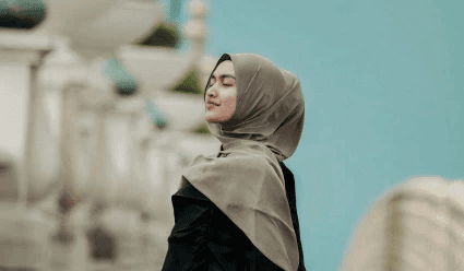 Young woman in a hijab standing outside with eyes closed and smiling.