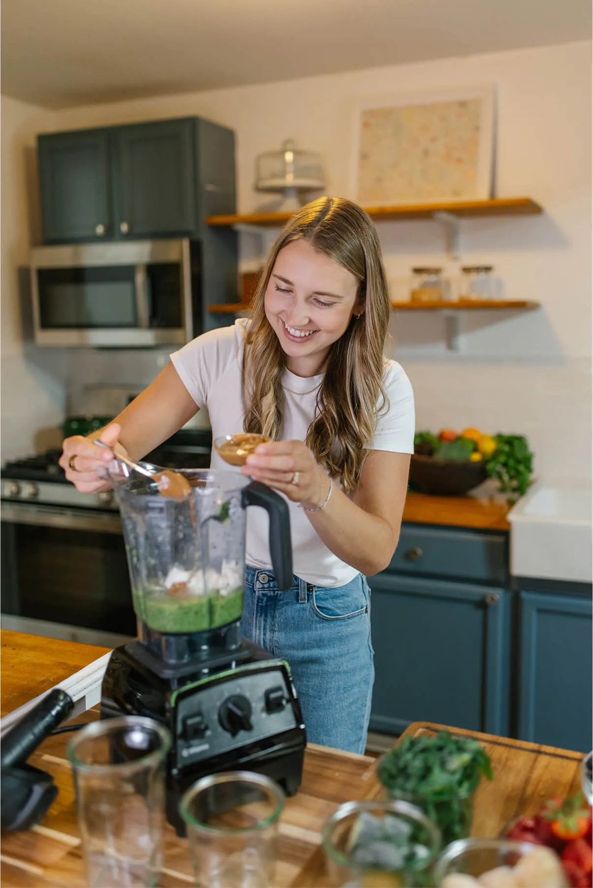 girl making a smoothie in a vitamix with greens, banana, protein powder, almond butter, in a kitchen that is clean and organized