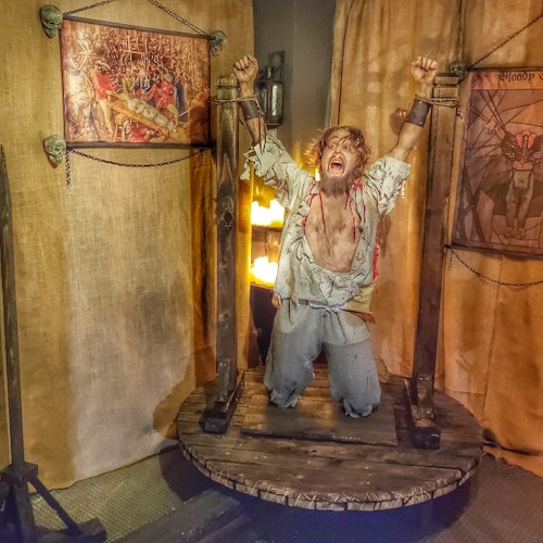 ایک آدمی پھٹے پرانے کپڑوں میں ایک لکڑی کے پلیٹ فارم سے جکڑا ہوا ہے، جو وسطی دور کے سجاوٹ اور تصاویر کے درمیان ہے، پریشان نظر آ رہا ہے۔