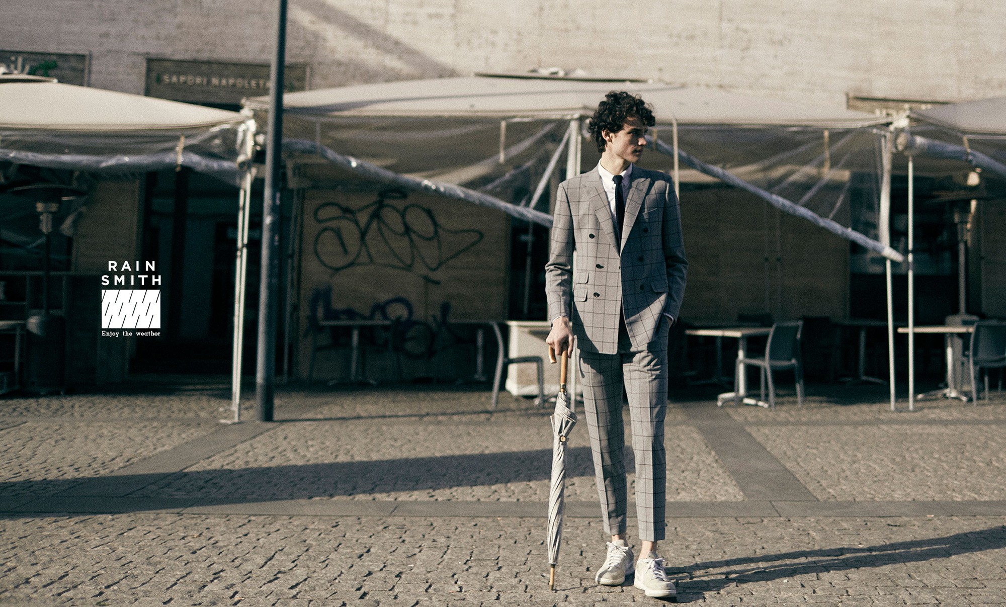這張圖像展示了 RainSmith 的形象攝影圖,一位穿著格紋西裝的男子站在戶外,手持一把雨傘,左側有 RainSmith 的品牌識別和 "Enjoy the weather" 字樣。