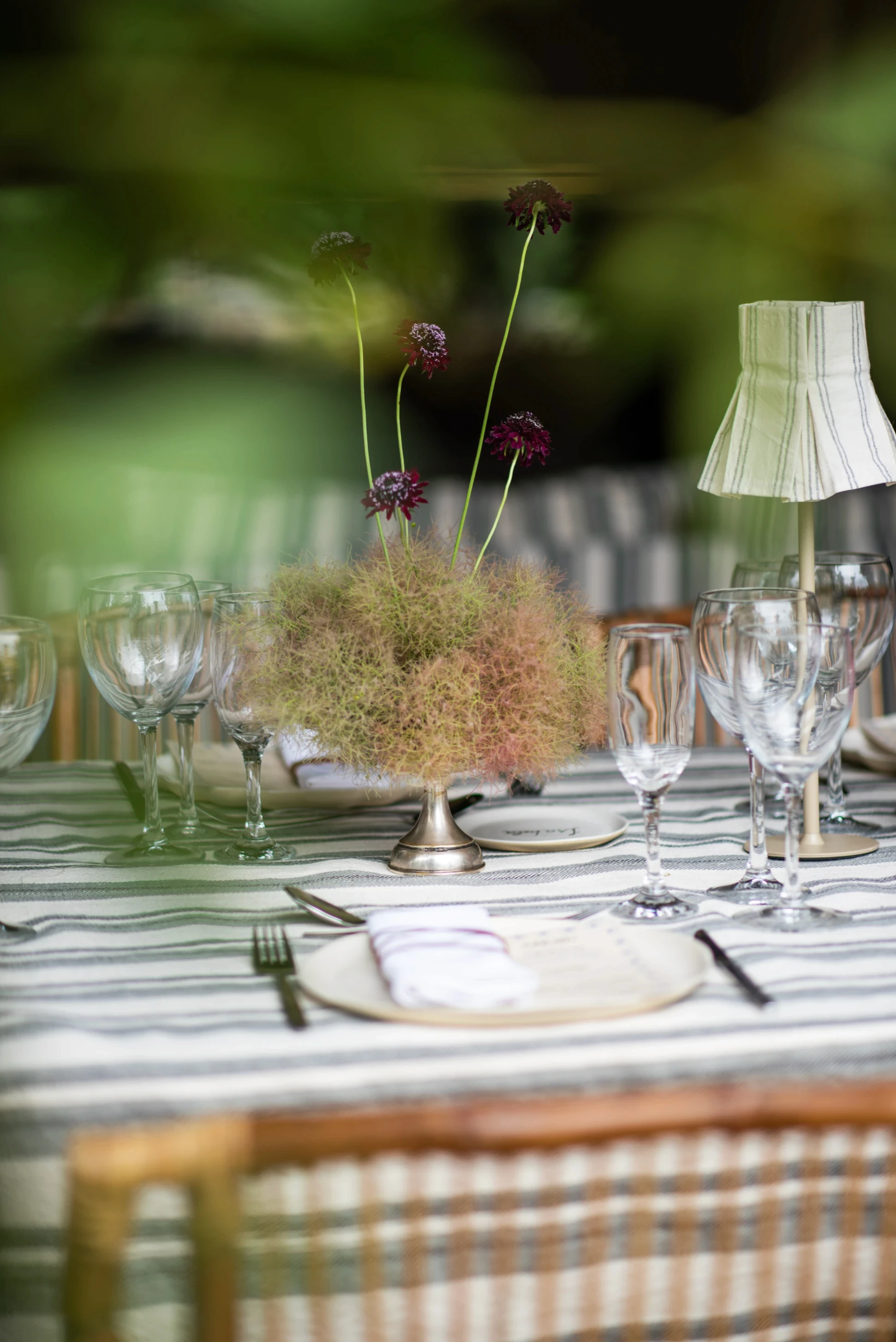 Puesta de mesa con centro etéreo de smoke bush y flores burdeos, copas de vino y mantelería a rayas neutras.
