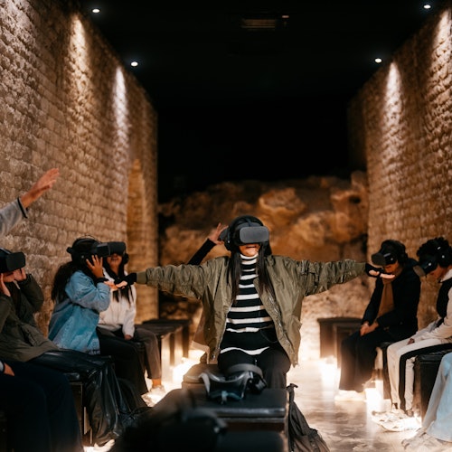 Một nhóm người đang đội thiết bị thực tế ảo ngồi trên các băng ghế trong một căn phòng có tường đá, một số với cánh tay duỗi ra, trải nghiệm thực tế ảo.