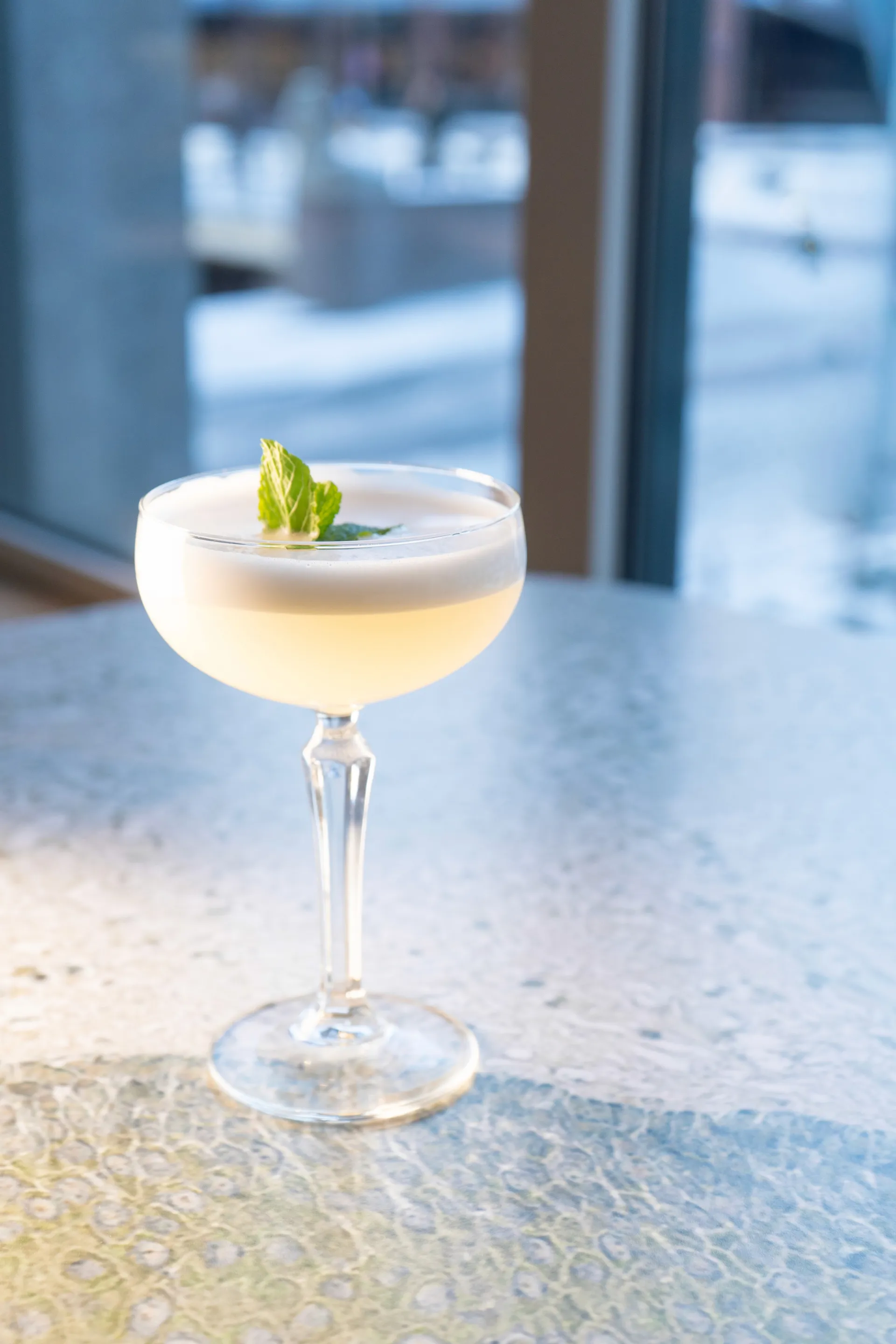 A elegantly garnished cocktail in a coupe glass sits on a table, with a blurred cityscape in the background.