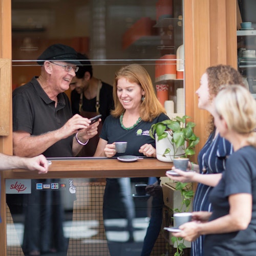 四个人在咖啡馆外享受咖啡，微笑并交谈。一名男子向柜台后面的女士展示手机。