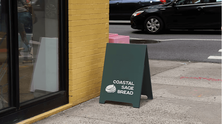 kelp green sandwich board with the coastal sage bread logo at the base in front of a yellow building 
