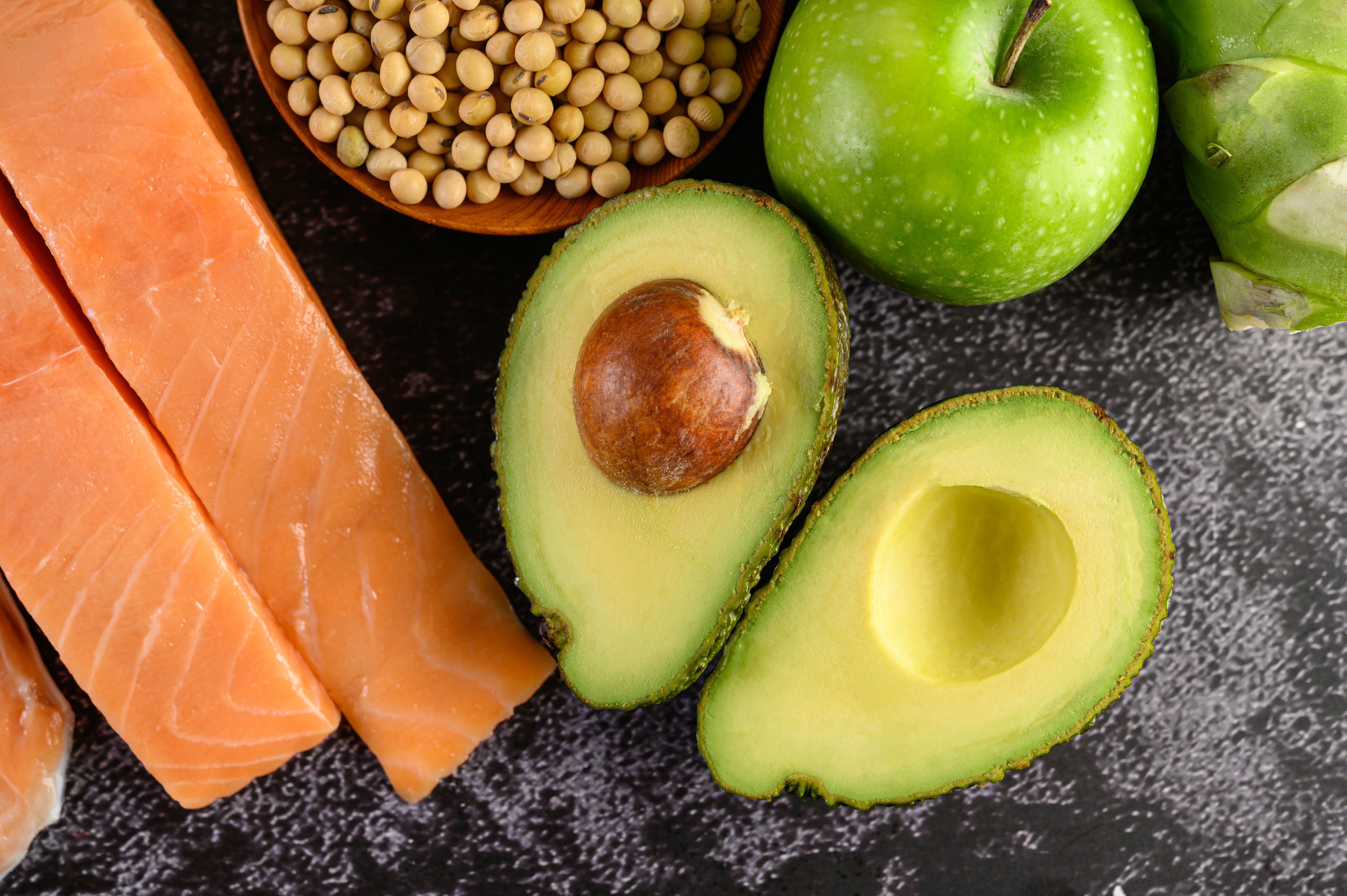 Close-up of fresh foods on a dark surface: salmon fillets, halved avocado, green apples, artichoke, and a bowl of soybeans, conveying health.