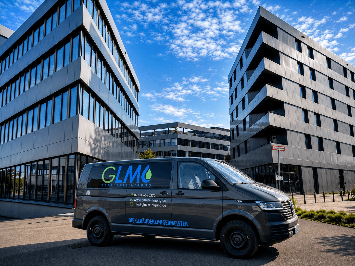 Ein grauer Transporter (Volkswagen) mit der Aufschrift „GLM GEBÄUDEREINIGUNG“ und dem Slogan „DIE GEBÄUDEREINIGERMEISTER“ steht auf einer asphaltierten Fläche vor einer modernen Bürolandschaft. Auf der Fahrzeugtür sind zudem Kontaktdaten wie Telefonnummer, Website und E-Mail-Adresse abgebildet.  Im Hintergrund ragen zwei markante, mehrstöckige Bürogebäude mit futuristisch-kantiger Architektur, metallisch glänzenden Fassadenplatten und großen Fensterfronten in den leicht bewölkten, blauen Himmel empor.
