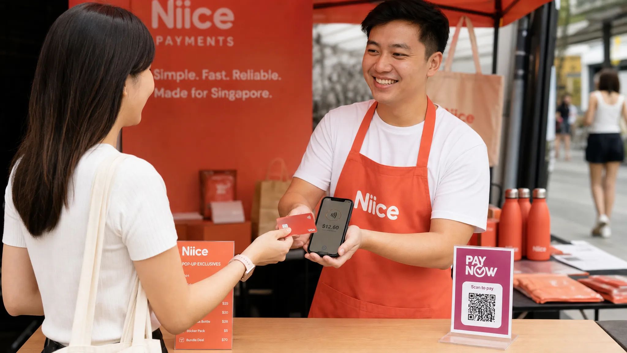 A Singapore pop-up stall where a customer taps a contactless card on a merchant's smartphone to pay, with a small PayNow QR sign also visible on the counter.