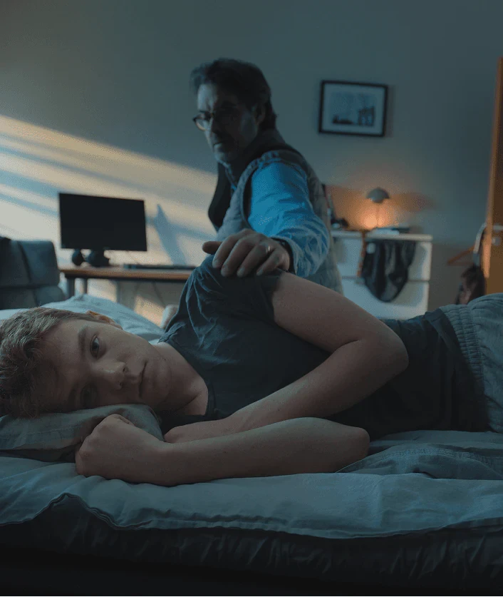 En pappa lägger handen på axeln till en tonåring som ligger vaken i sängen