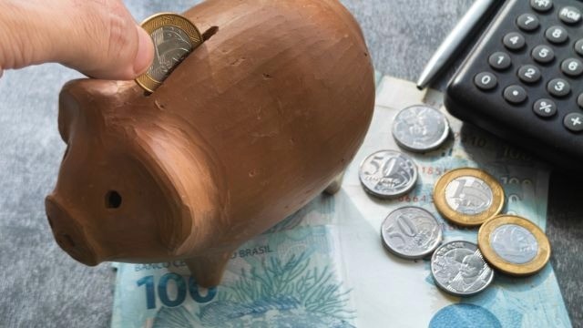 Cofre de moedas em formato de porquinho sobre notas e moedas de real próximo a uma calculadora e caneta