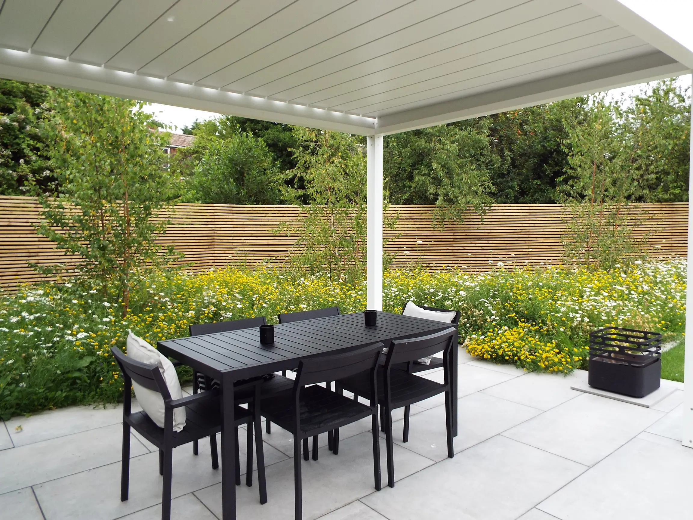 A shaded patio area with a black table and chairs, surrounded by lush greenery and a fence in the background.