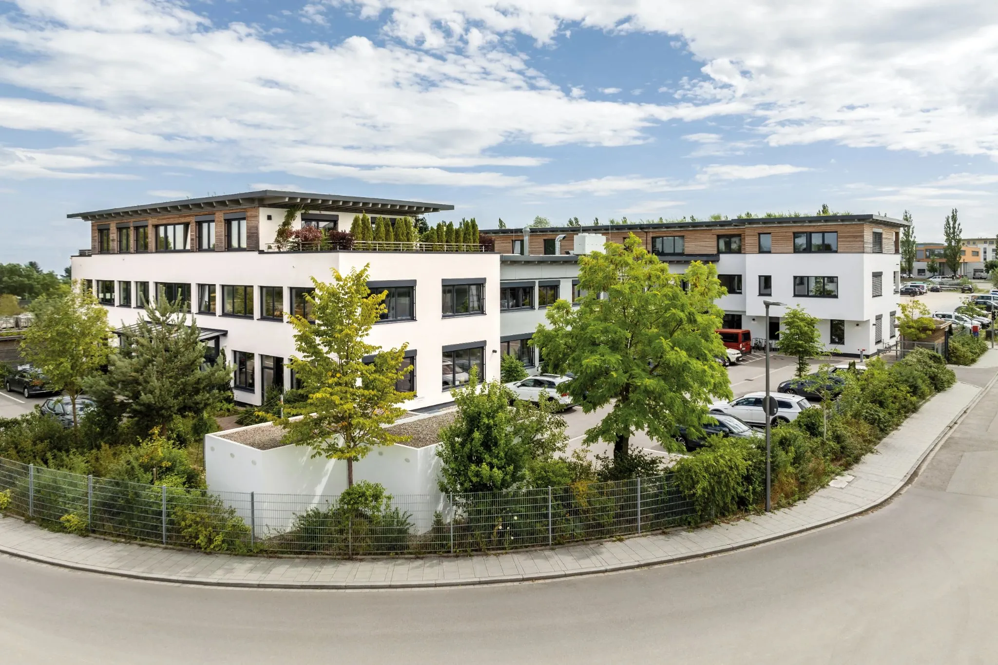Moderne Bürogebäude mit begrünten Dächern und viel Grünfläche, geparkte Fahrzeuge und ein bewölkter Himmel bei Tageslicht.