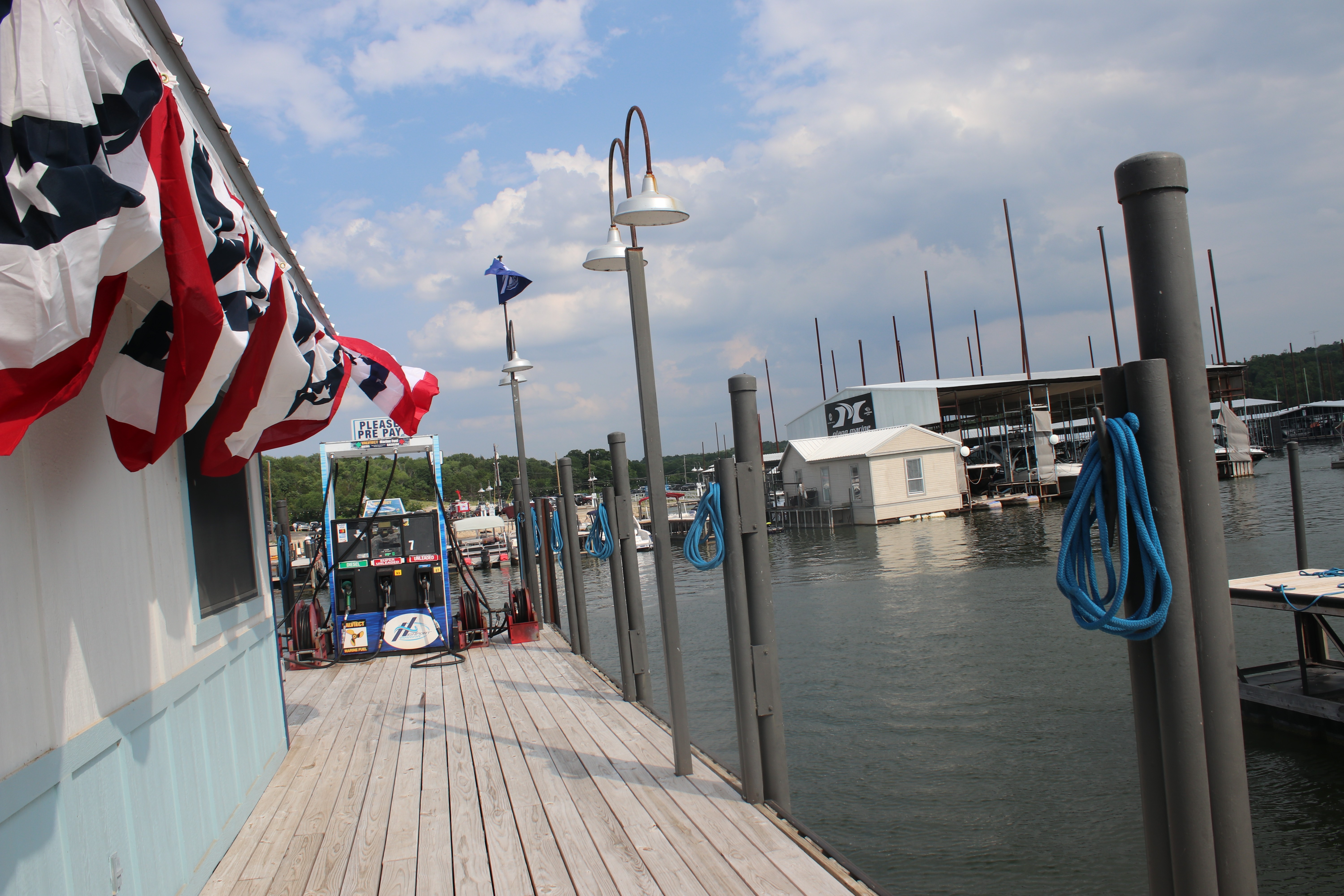 A lakeside marina lined with festive red, white, and blue banners features a wooden boardwalk leading to moored boats under a partly cloudy sky.
