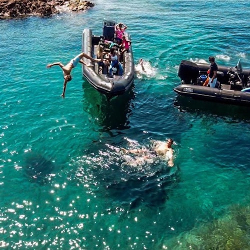 Folk på oppblåsbare båter med én person som dykker ned i klart, blått vann, mens andre svømmer i nærheten. Steiner er synlige i bakgrunnen.