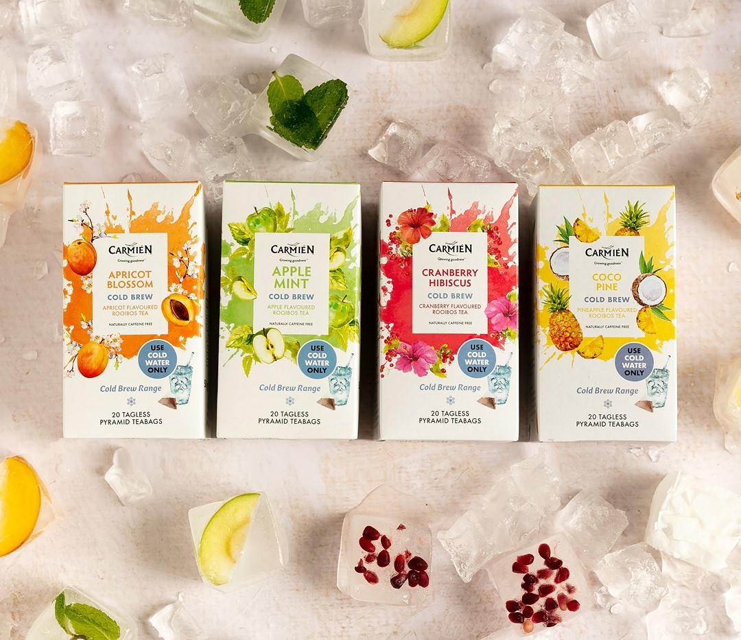 Four boxes of different Carmién tea blends laying flat on a white counter surrounded by ice blocks and various pieces of fruit.