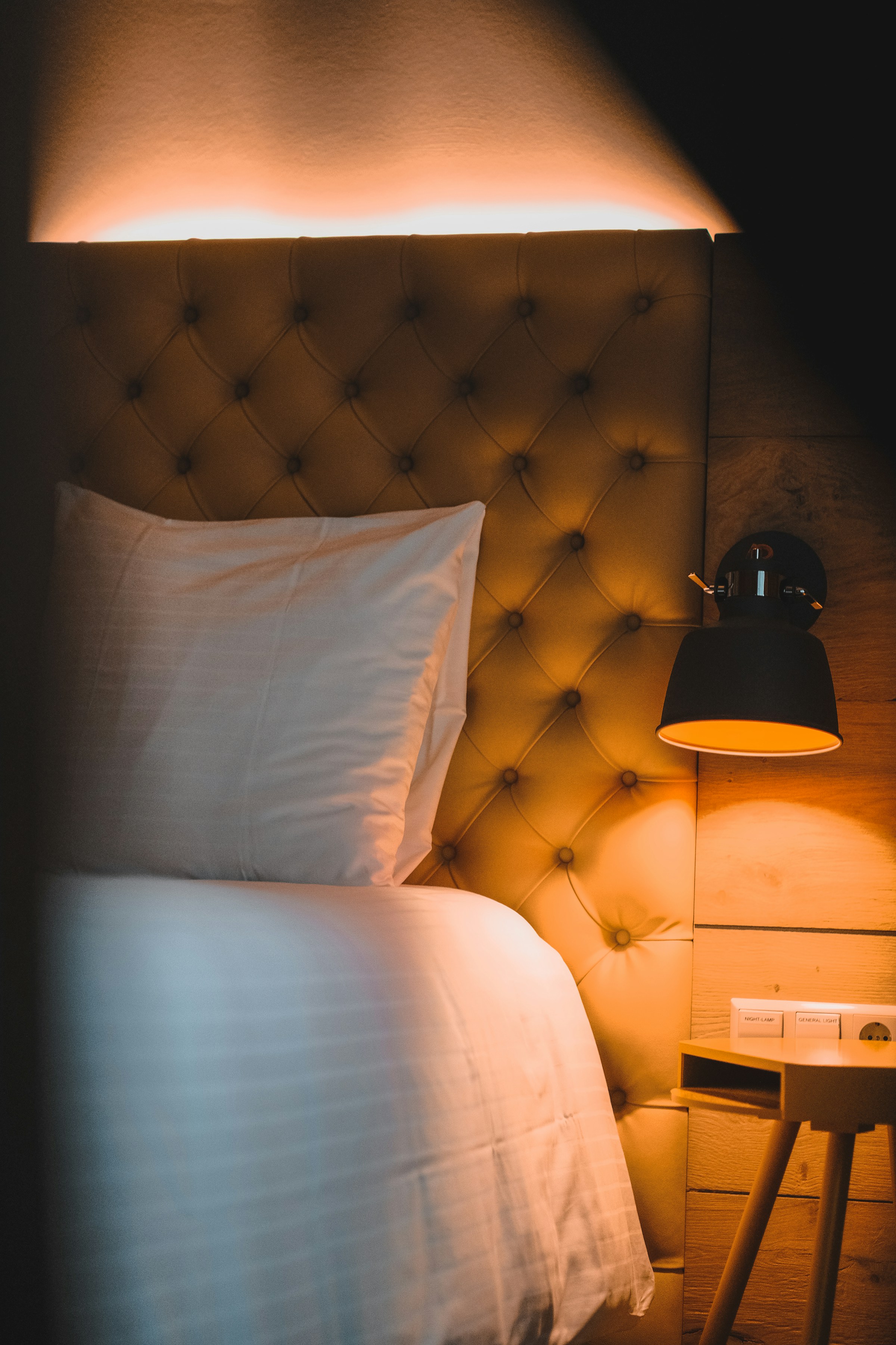 A lamp-lit upholstered headboard and white bed linen in a dim room