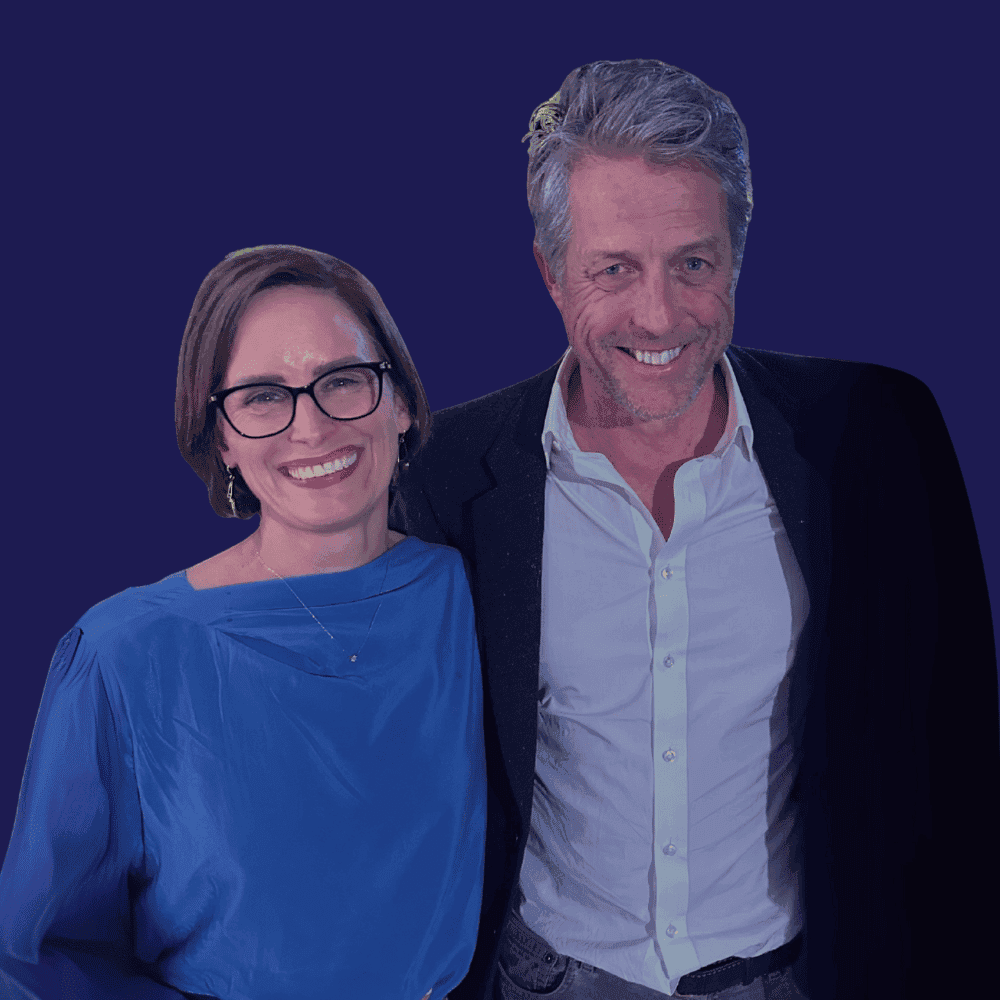 A smiling woman in a blue blouse and glasses stands beside a man in a dark suit and white shirt, set against a plain dark background.