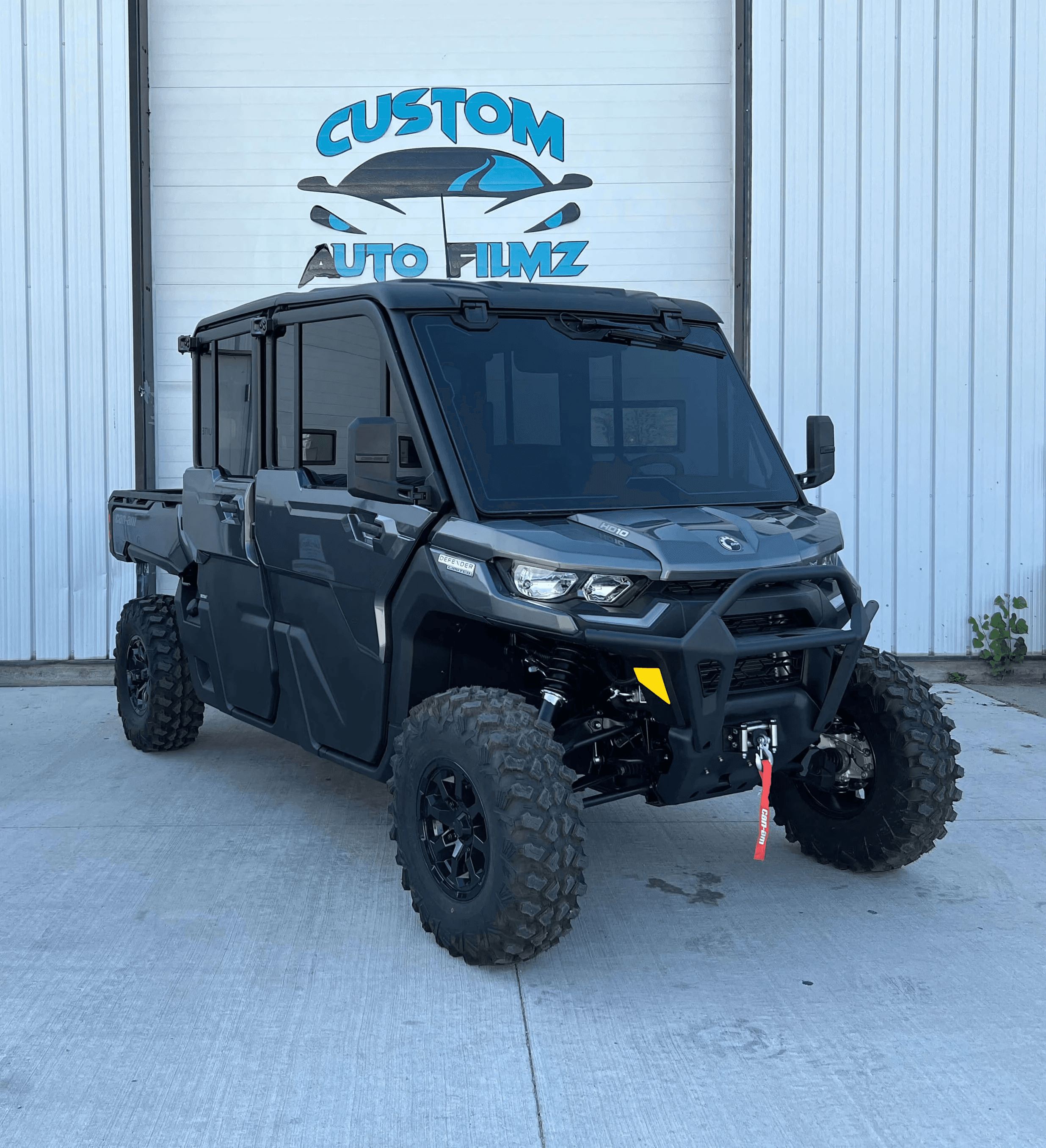 A black Can-Am Defender side-by-side utility vehicle is parked on a concrete driveway in front of a white industrial building.